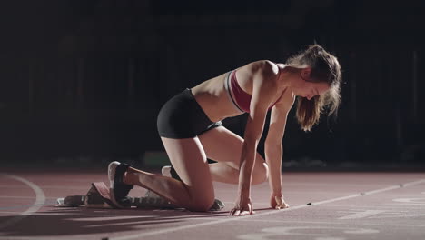 Die-Frau-Am-Start-Des-Rennens-Steigt-In-Die-Pads,-Erhebt-Sich-Und-Rennt-In-Zeitlupe-Abends-Im-Stadion.-Läuferin-Duckt-Sich-In-Die-Ausgangsposition,-Bevor-Sie-Mit-Dem-Rennen-Beginnt.